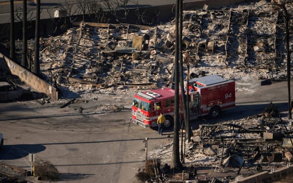 a picture of a bright red fire engine sitting in front of the shells of a number of burned out houses, all in grey rubble